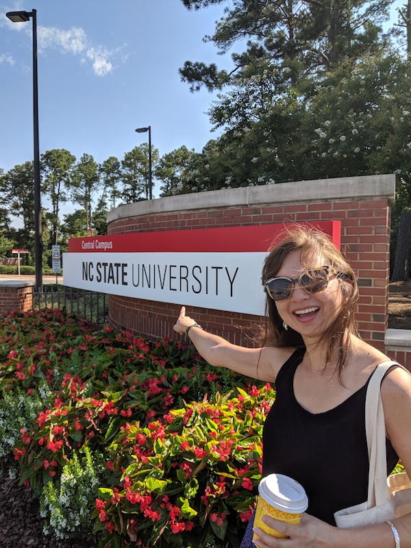 Robin smiling and pointing at an NC State University sign